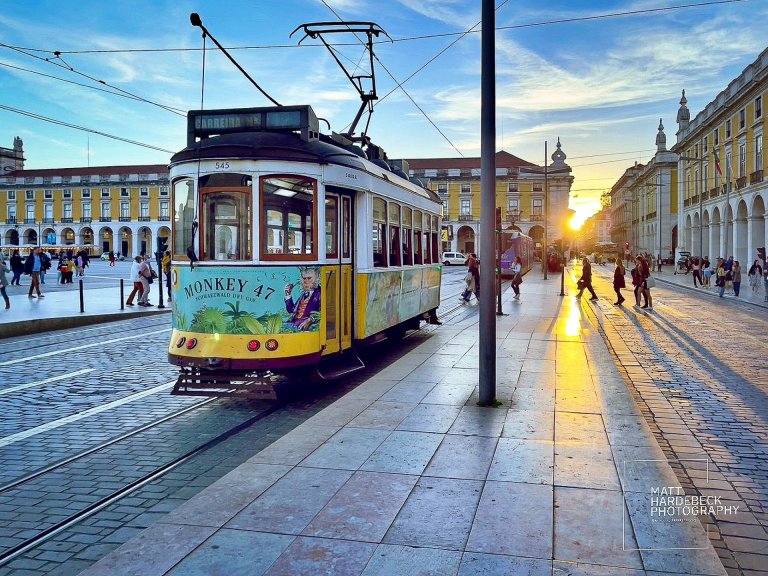 Historic yellow tram in Lisbon's Praça do Comércio at sunset, with long shadows stretching across the cobblestone street.