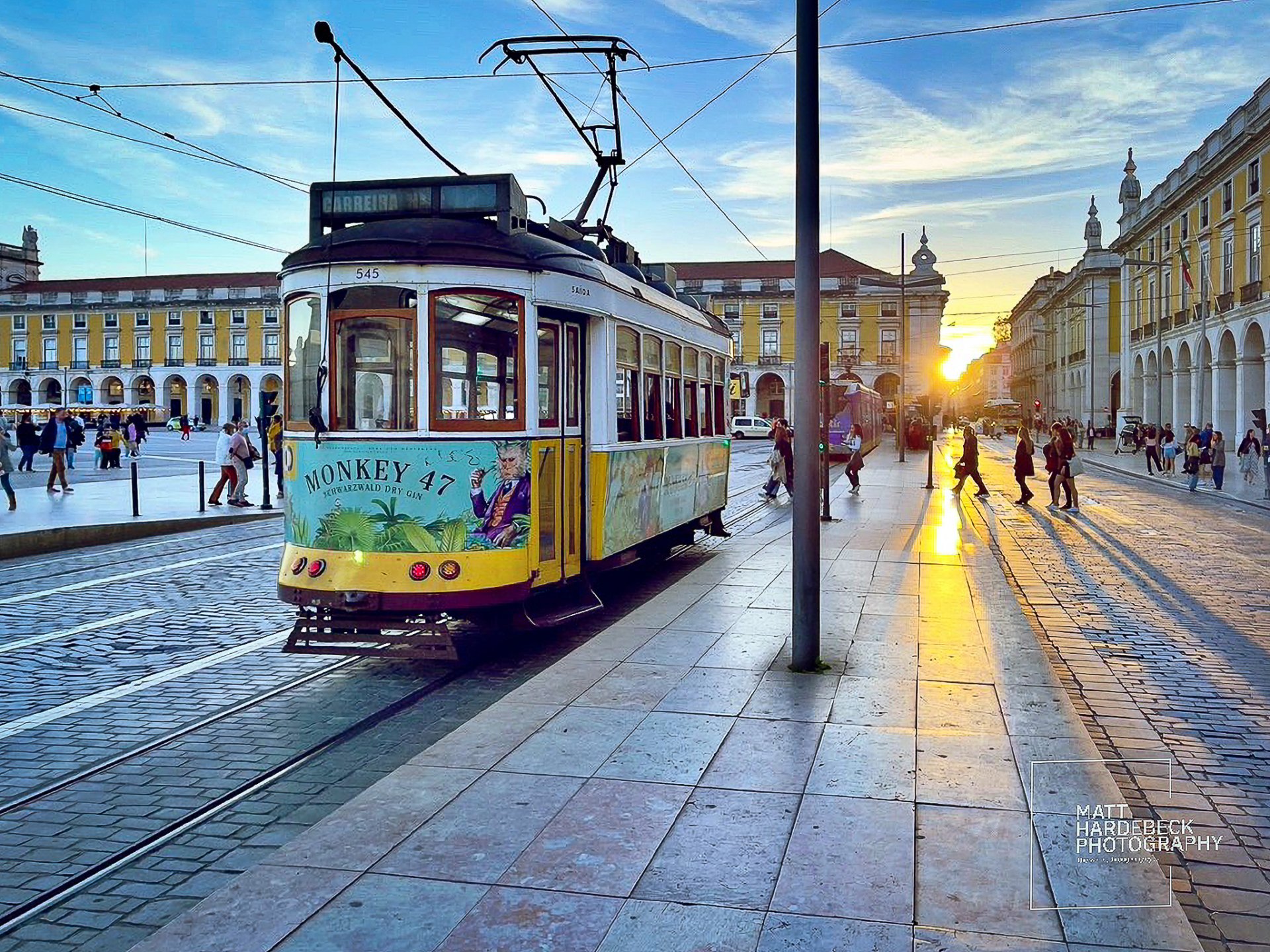 Historic yellow tram in Lisbon's Praça do Comércio at sunset, with long shadows stretching across the cobblestone street.