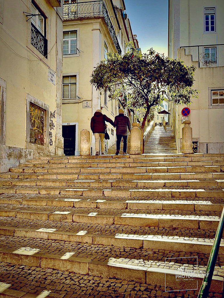 Two people walking up sunlit stone steps in Lisbon’s historic district, framed by a small tree.