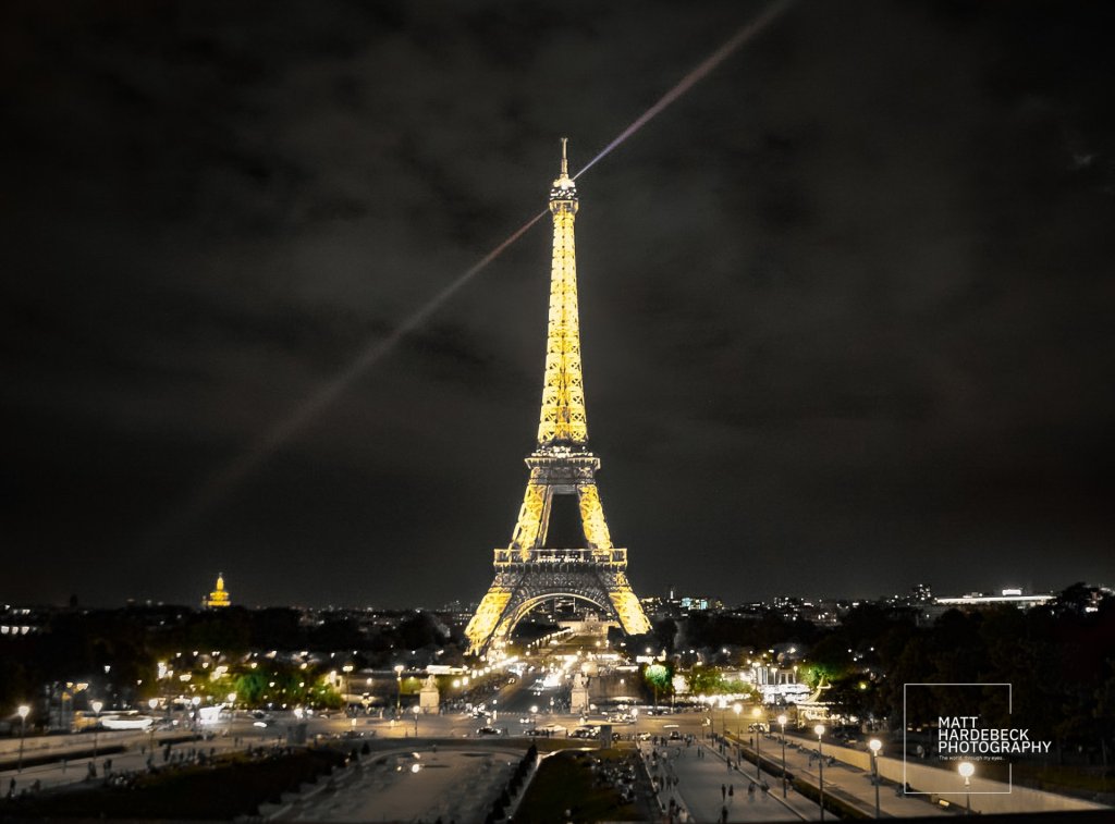 The Eiffel Tower illuminated at night, viewed from the Trocadéro with city lights glowing below and a spotlight beaming across the sky.