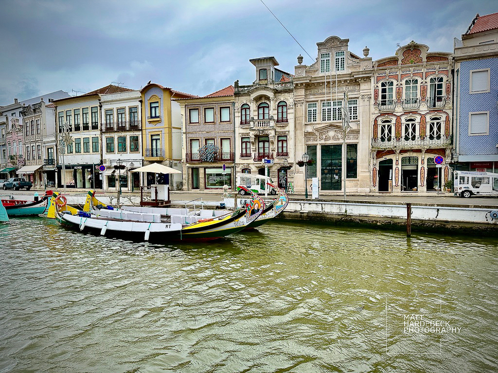 Moliceiro boats moored along the canal in Aveiro, Portugal, with ornate historic buildings in the background.
