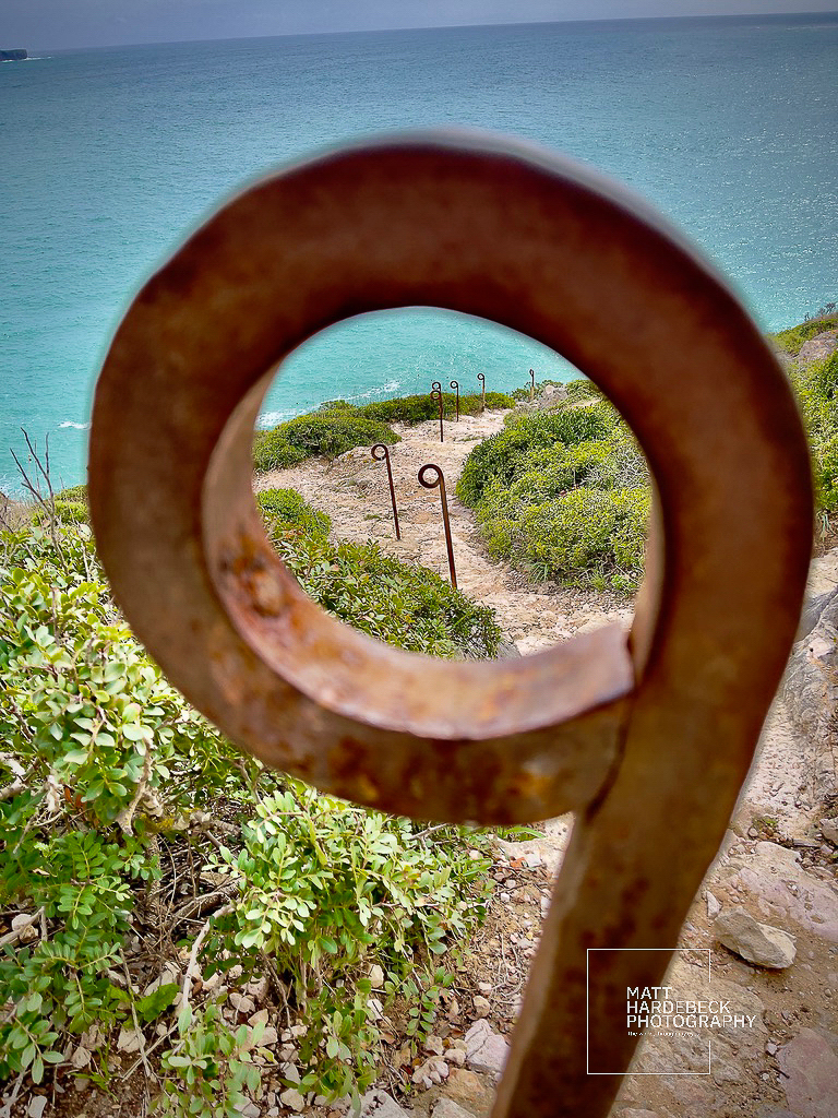 Rusted metal ring in the foreground framing a descending cliffside path to the ocean at Cabo de São Vicente, Portugal.