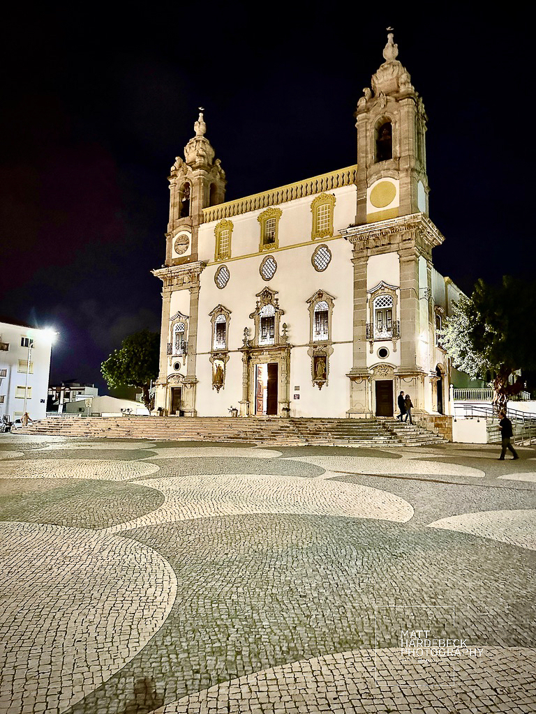 Illuminated Baroque church facade at night, fronted by a patterned cobblestone square.