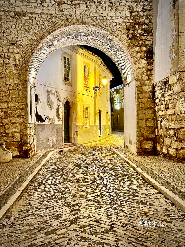 Stone archway leading to a cobblestone street lined with historic whitewashed buildings at night.