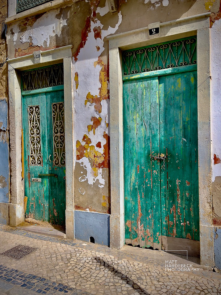 Two weathered teal doors on a peeling, sun-faded facade in Faro, Portugal.