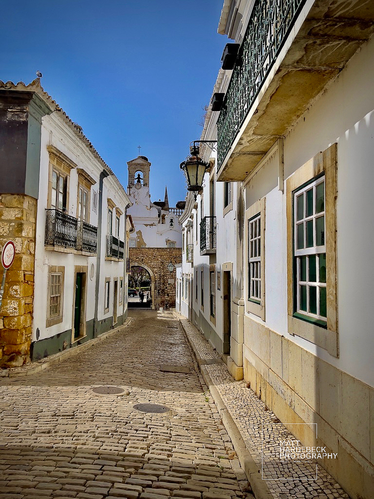 Narrow cobblestone street in Faro’s old town leading toward the Arco da Vila under a bright blue sky.