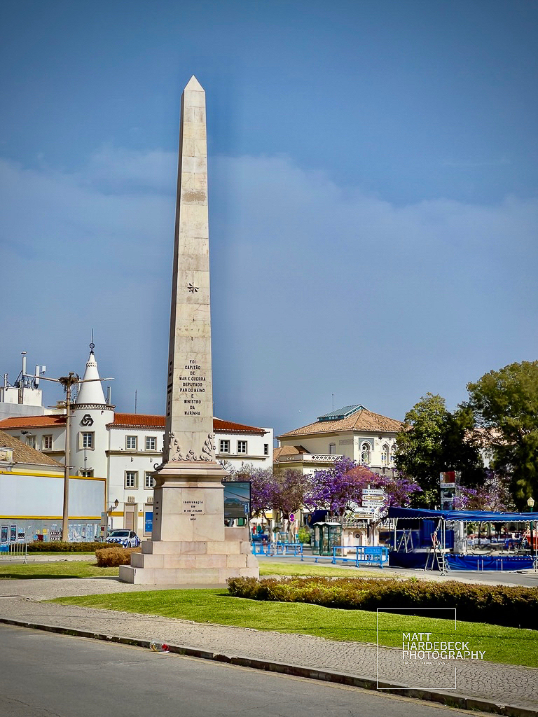 Tall stone obelisk monument in a landscaped square in Faro, Portugal.