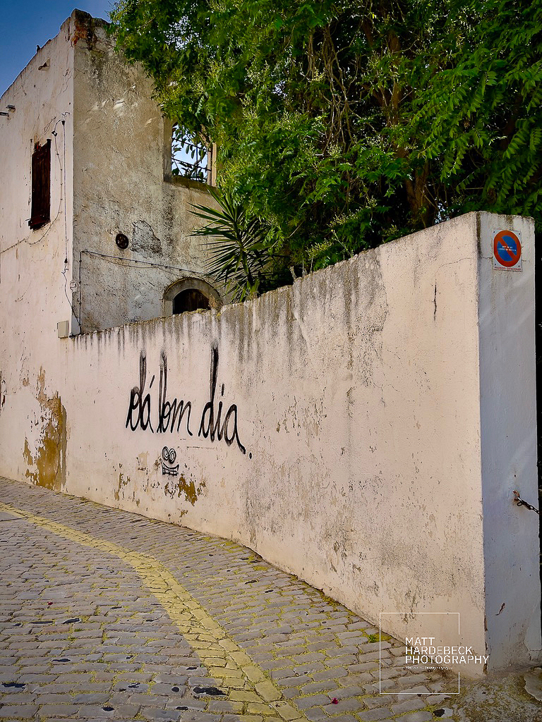 Graffiti reading “Até bem dia” on a weathered wall beside a cobblestone street in Faro.