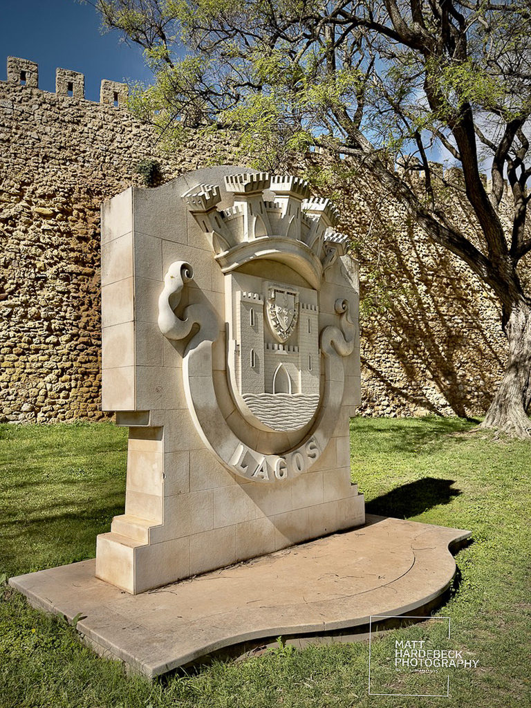 Stone crest of Lagos, Portugal, set against the historic city walls and shaded by a tree.