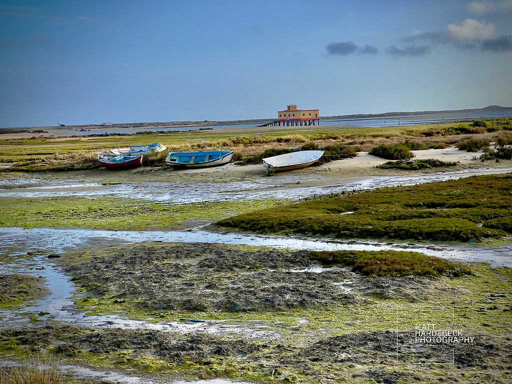 Row of small fishing boats resting on the shore in Fuseta with a stilted building in the distance.