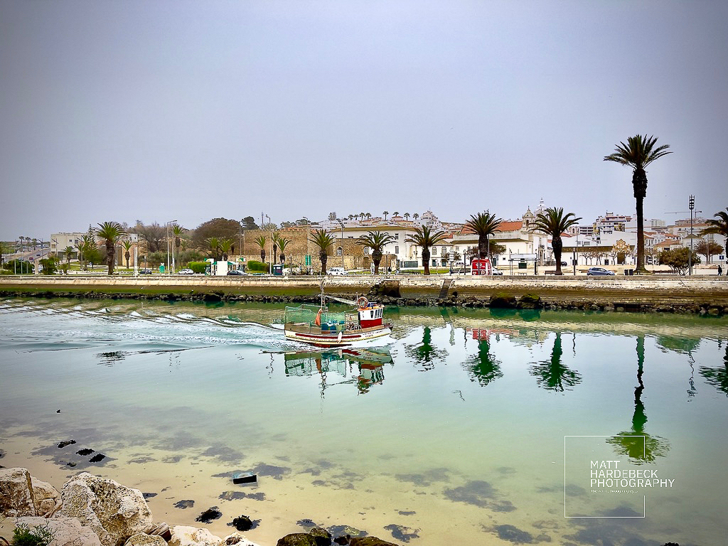 Small fishing boat crossing the Lagos channel, with palm-lined promenade behind.