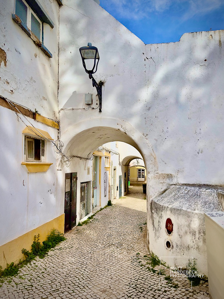 Narrow cobblestone alleyway in Lagos, Portugal, passing under a whitewashed arch with a black lantern.
