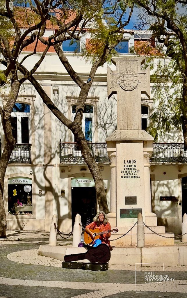 Street musician playing guitar beside a war memorial in Lagos, Portugal.