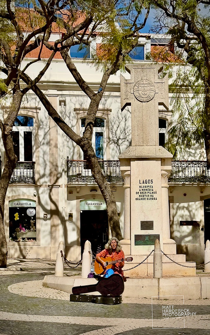 Street musician playing guitar beside a war memorial in Lagos, Portugal.