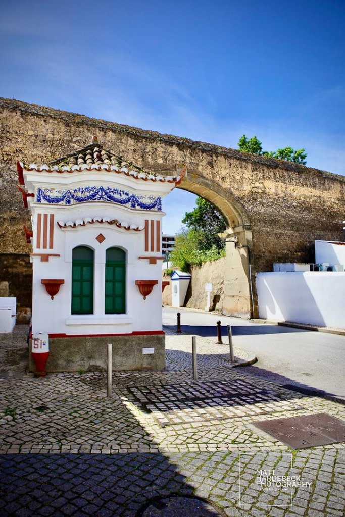 Small white building with green windows and blue tile trim in front of an ancient stone city gate in Lagos, Portugal.