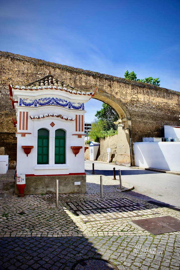 Small white building with green windows and blue tile trim in front of an ancient stone city gate in Lagos, Portugal.