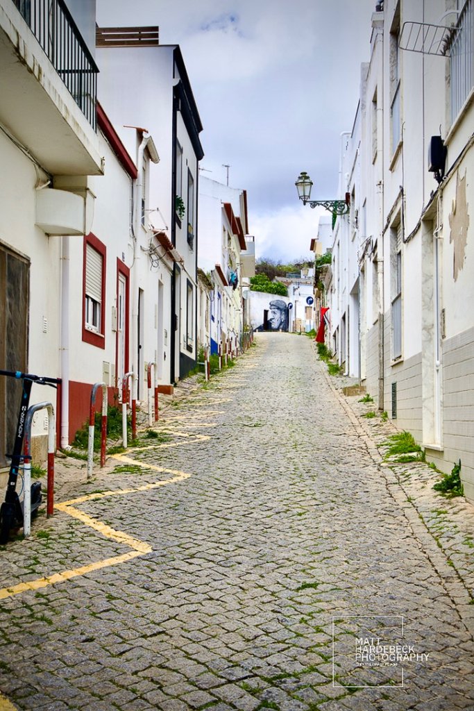 Steep cobblestone street lined with whitewashed buildings leading to a mural in Lagos, Portugal.