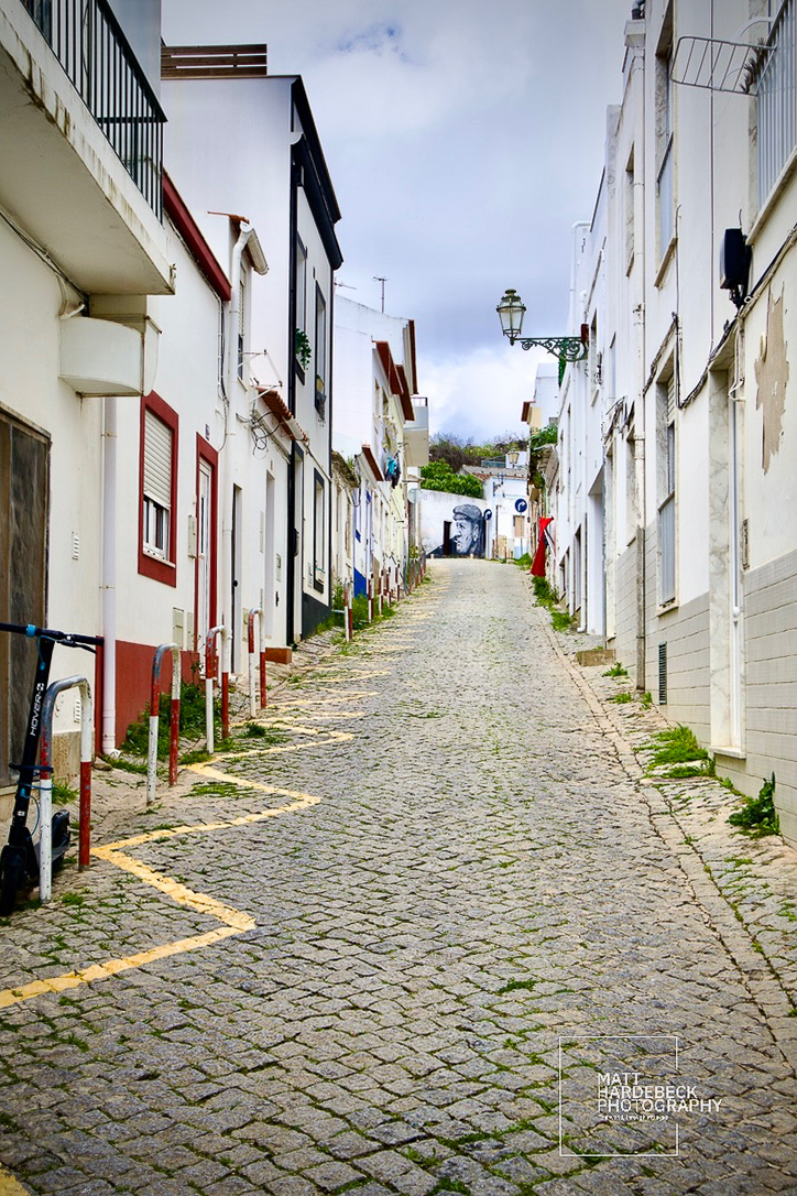 Steep cobblestone street lined with whitewashed buildings leading to a mural in Lagos, Portugal.