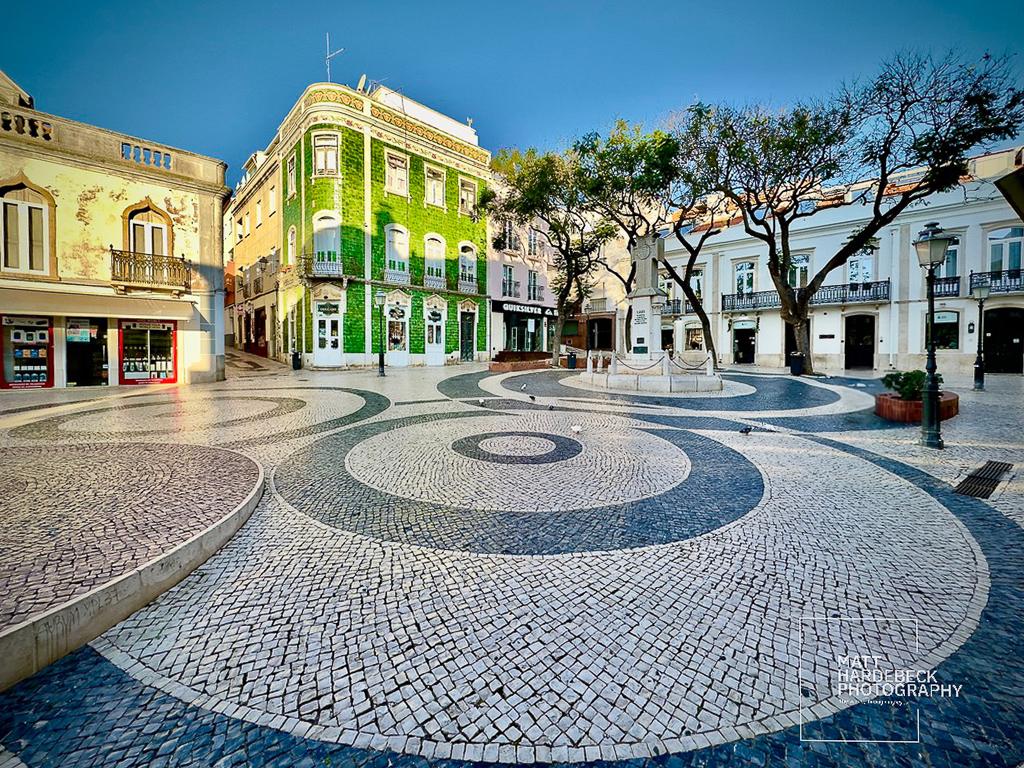 Open plaza with black-and-white wave-patterned pavement and green-tiled building in Lagos, Portugal.