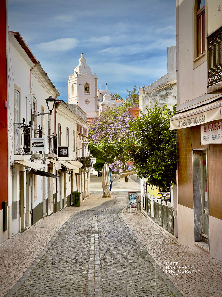 View down a cobblestone street in Lagos, Portugal, with whitewashed buildings and a church bell tower in the distance.