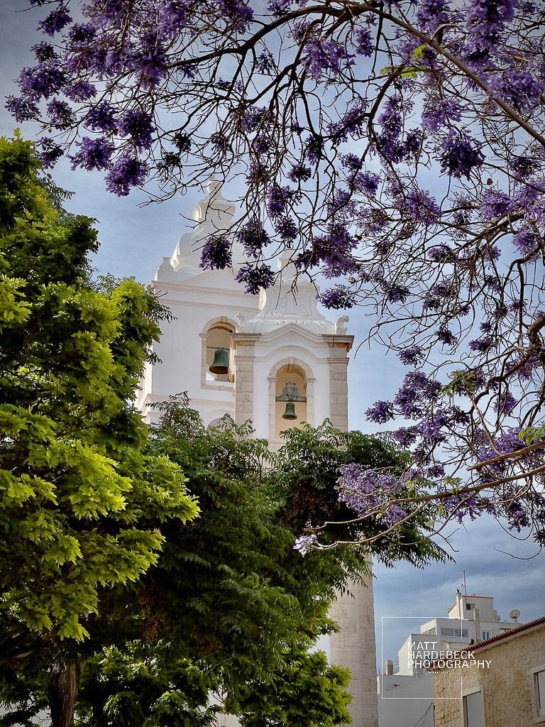 White church bell tower in Lagos, Portugal, framed by purple jacaranda blossoms.