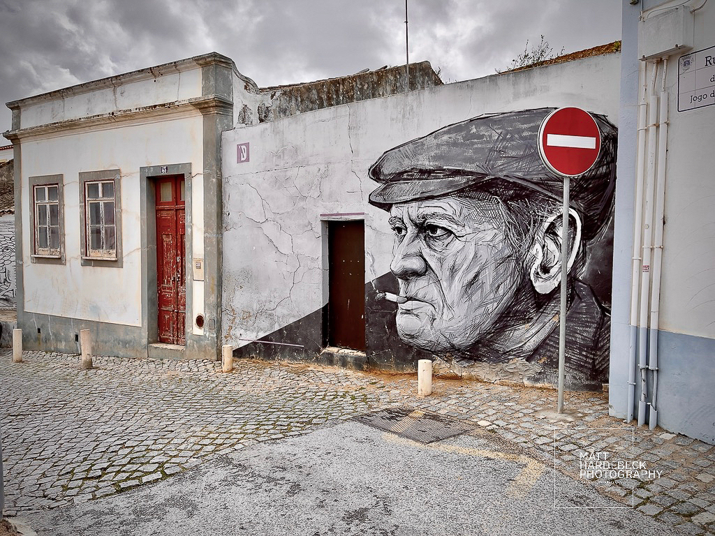 Street art mural of an elderly man in a cap smoking, painted on the side of a weathered building in Lagos, Portugal.