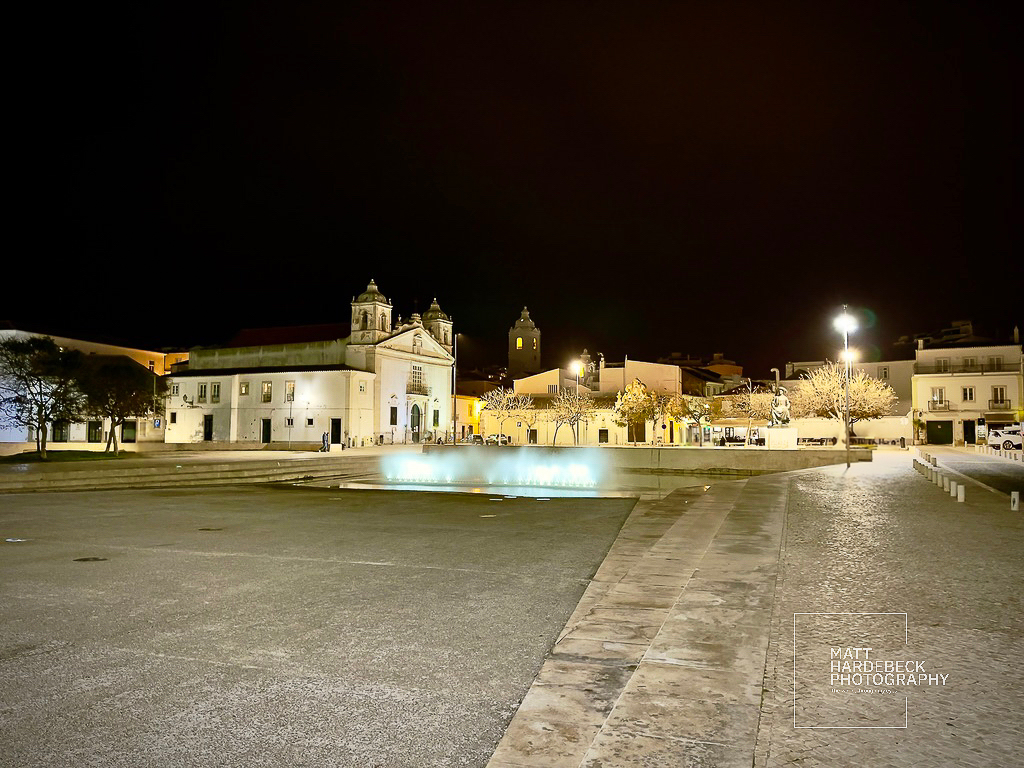 Night view of a square in Lagos, Portugal, featuring a church, illuminated buildings, and a central fountain.