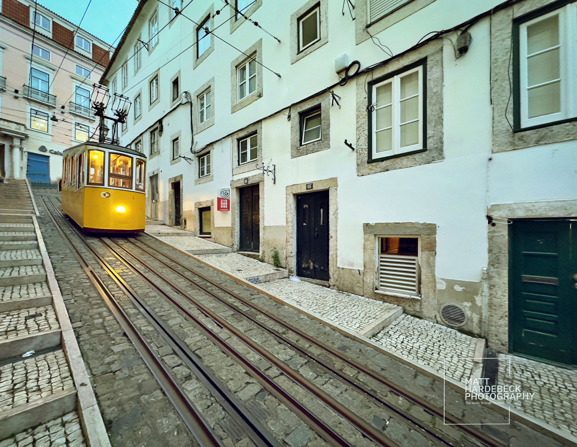 A yellow funicular car climbs a cobblestone street between whitewashed buildings in Lisbon.