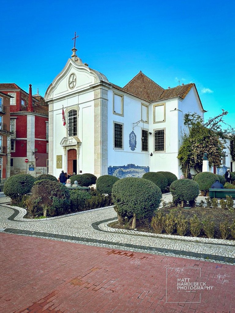 White church with a cross, manicured gardens, and patterned pavement in Lisbon.