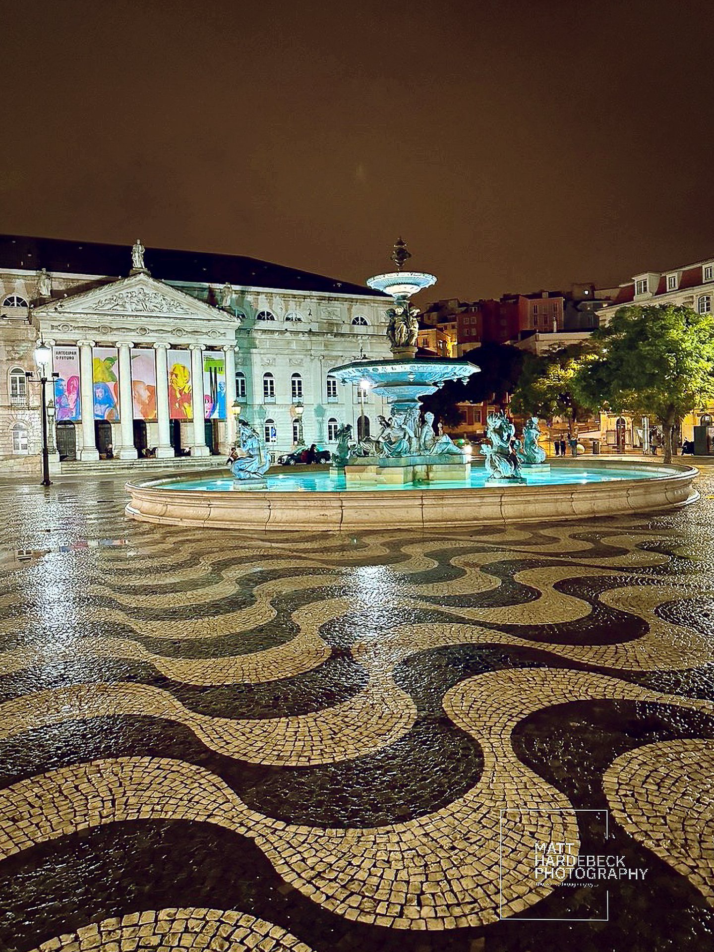 Night view of Rossio Square’s wave-patterned cobblestones with illuminated fountain and the neoclassical National Theatre D. Maria II in Lisbon, Portugal.