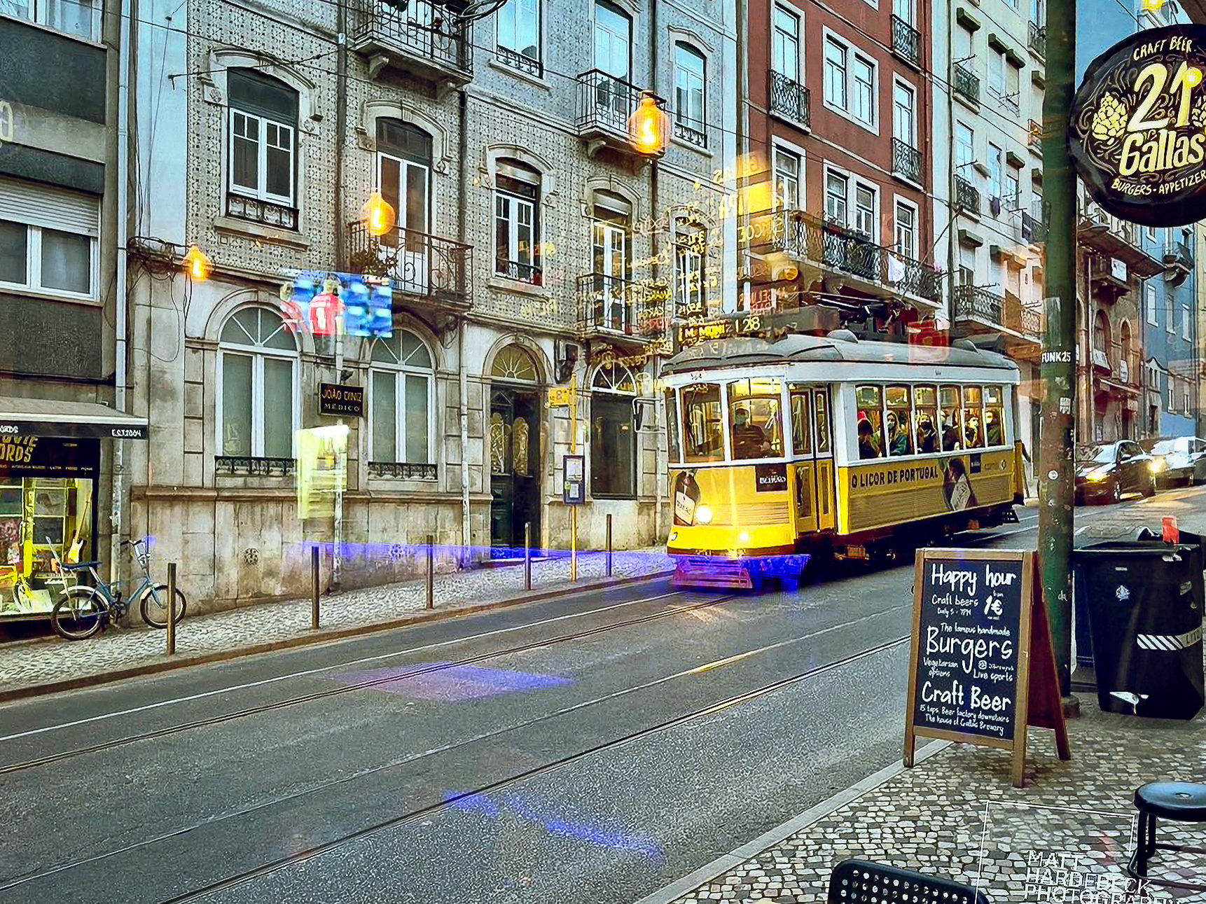 Iconic yellow Tram 28 passing through a Lisbon street lined with traditional tiled buildings, seen from a café window.