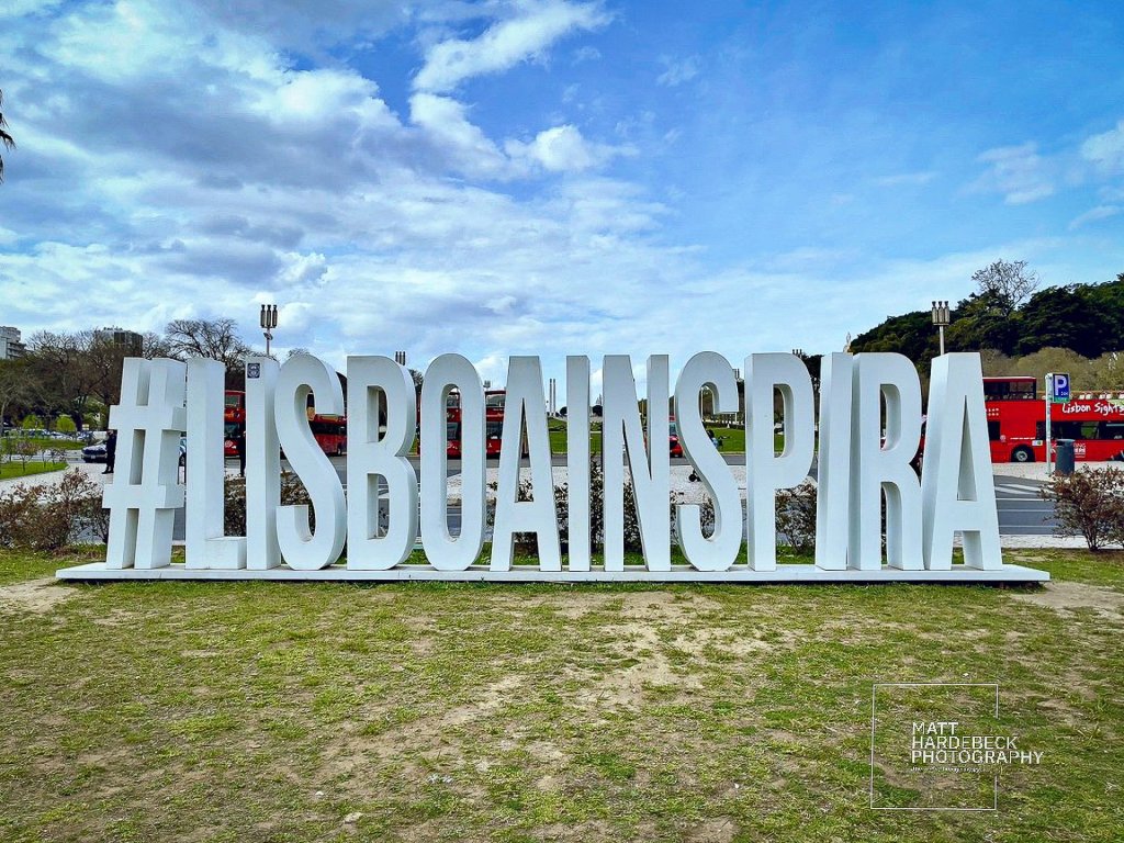 Large white sculpture spelling “#LISBOAINSPIRA” in a park in Lisbon, Portugal, with sightseeing buses and a partly cloudy sky in the background.