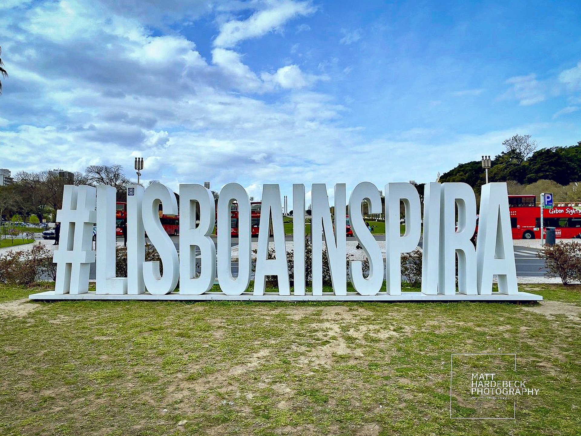 Large white sculpture spelling “#LISBOAINSPIRA” in a park in Lisbon, Portugal, with sightseeing buses and a partly cloudy sky in the background.