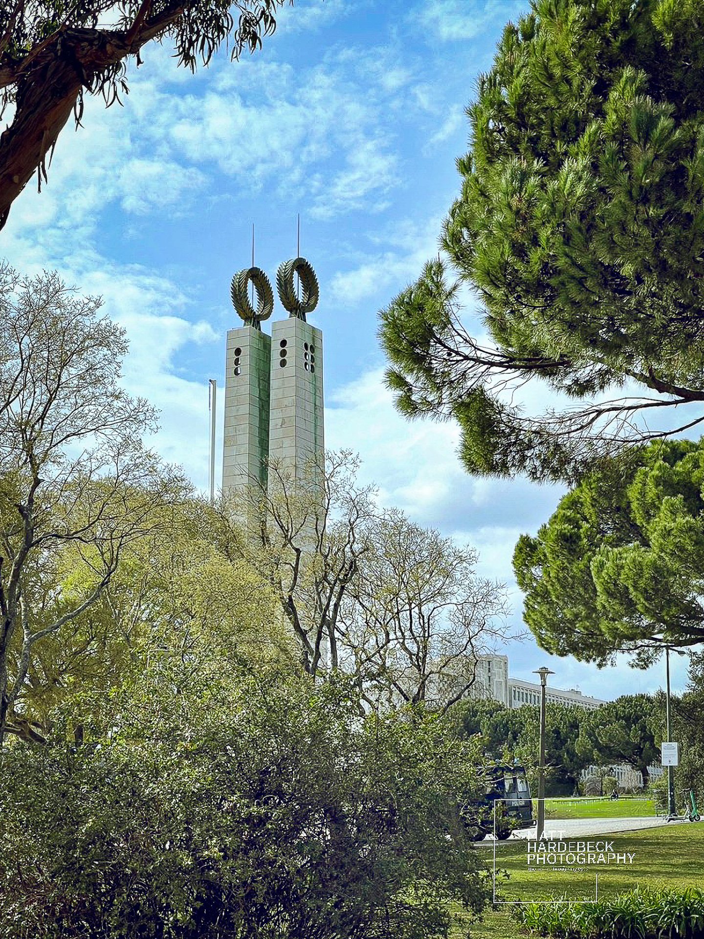 Twin stone towers with laurel wreath sculptures at Eduardo VII Park in Lisbon, framed by leafy trees under a blue sky.
