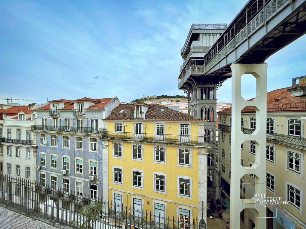 The ornate iron structure of the Santa Justa Lift connecting Lisbon’s Baixa district to Carmo Square, with colorful tiled buildings in the foreground.