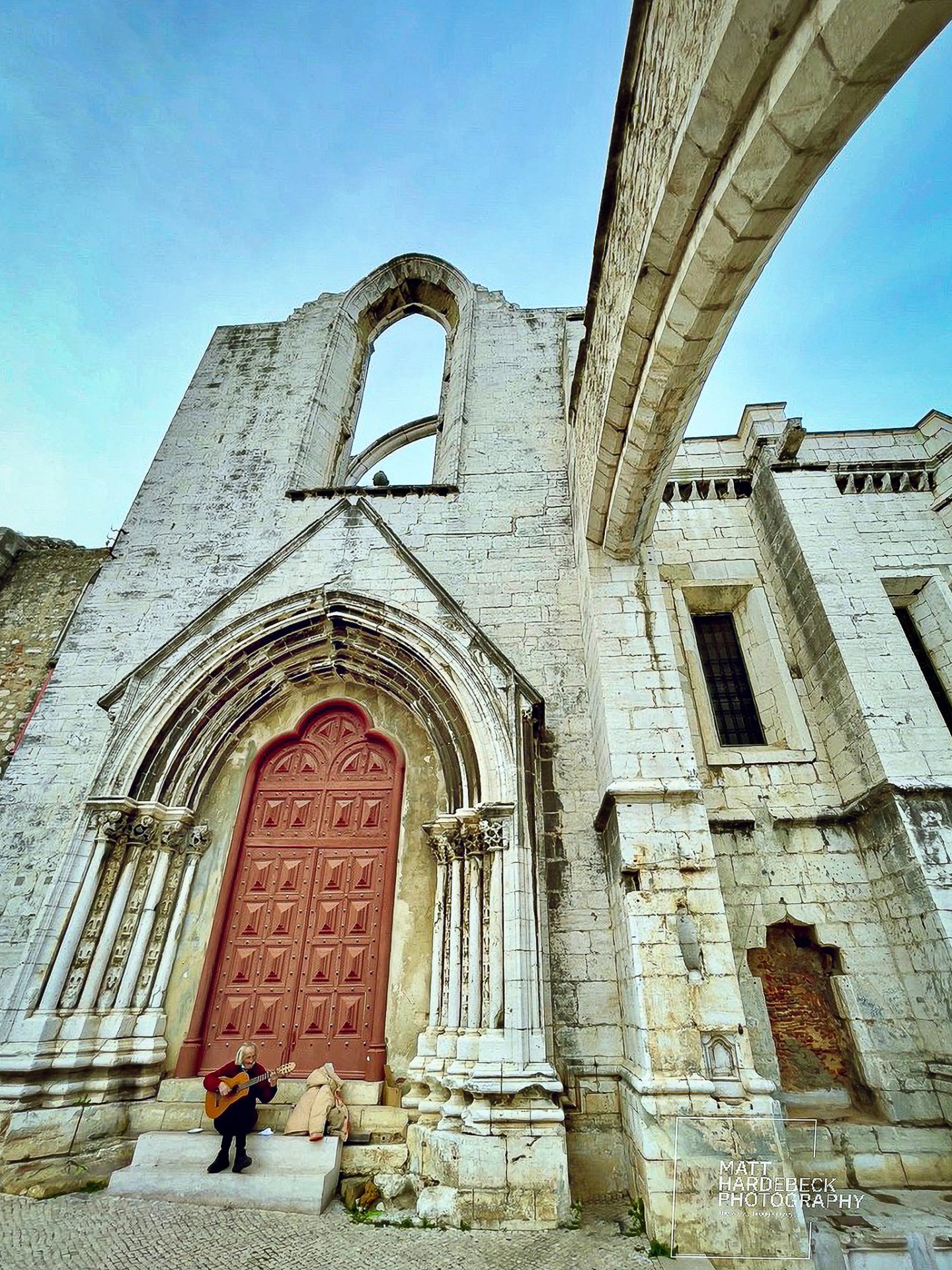 The Gothic entrance of the Carmo Convent in Lisbon with a musician playing guitar at its base.