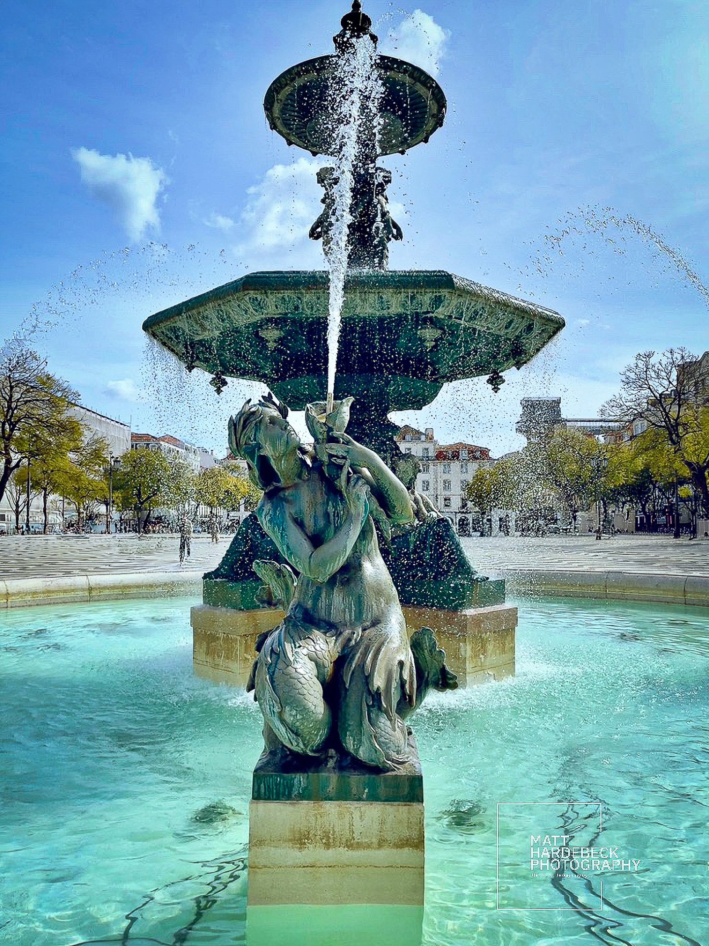 Bronze fountain in Lisbon’s Rossio Square with a statue of a sea nymph, water spraying under a clear sky.