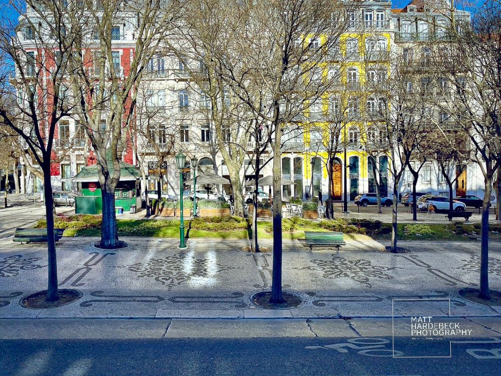 Tree-lined promenade in Lisbon with patterned Portuguese pavement and colorful 19th-century buildings in the background.