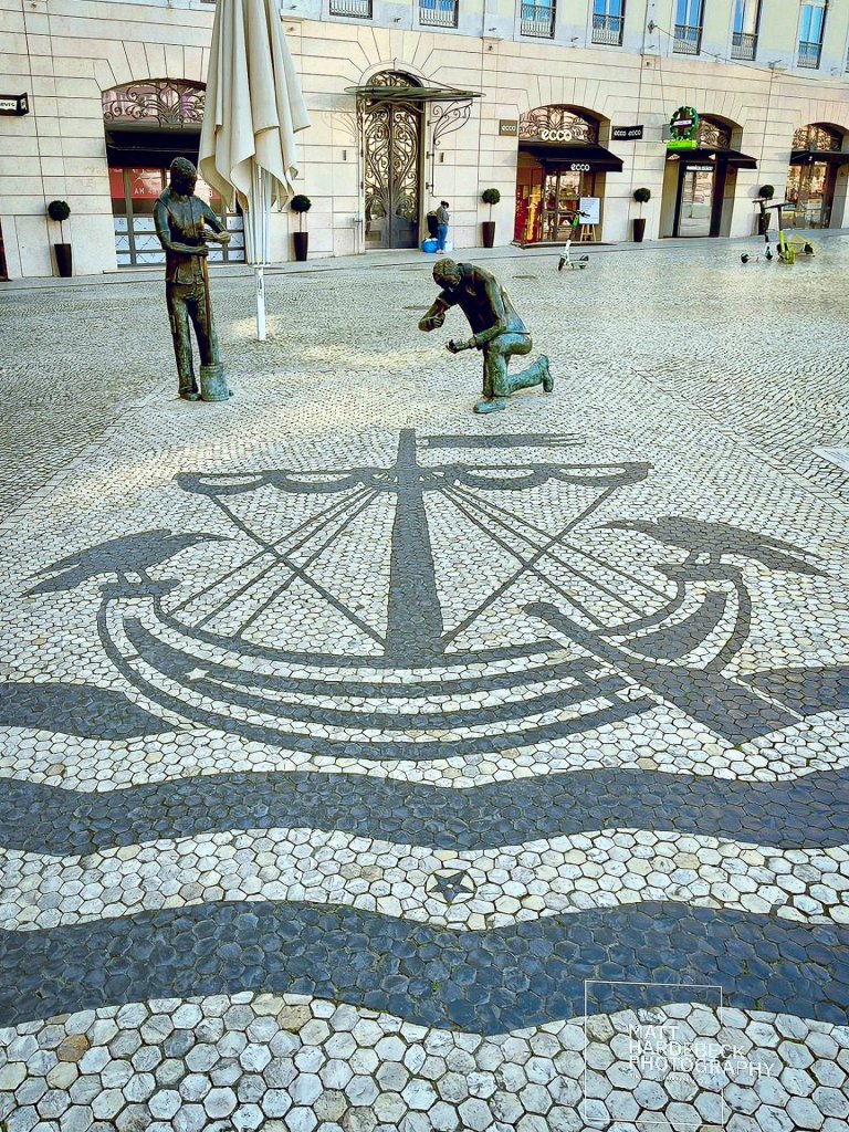 Black-and-white mosaic pavement depicting a ship and waves, flanked by bronze statues in Lisbon, Portugal.