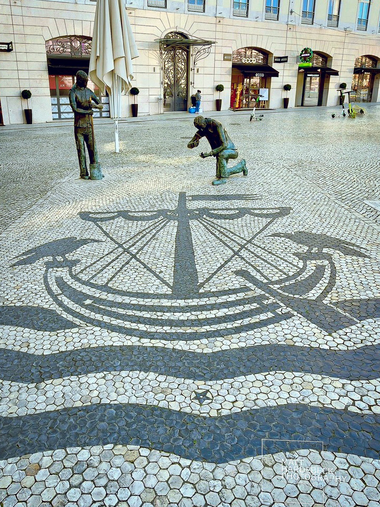 Black-and-white mosaic pavement depicting a ship and waves, flanked by bronze statues in Lisbon, Portugal.