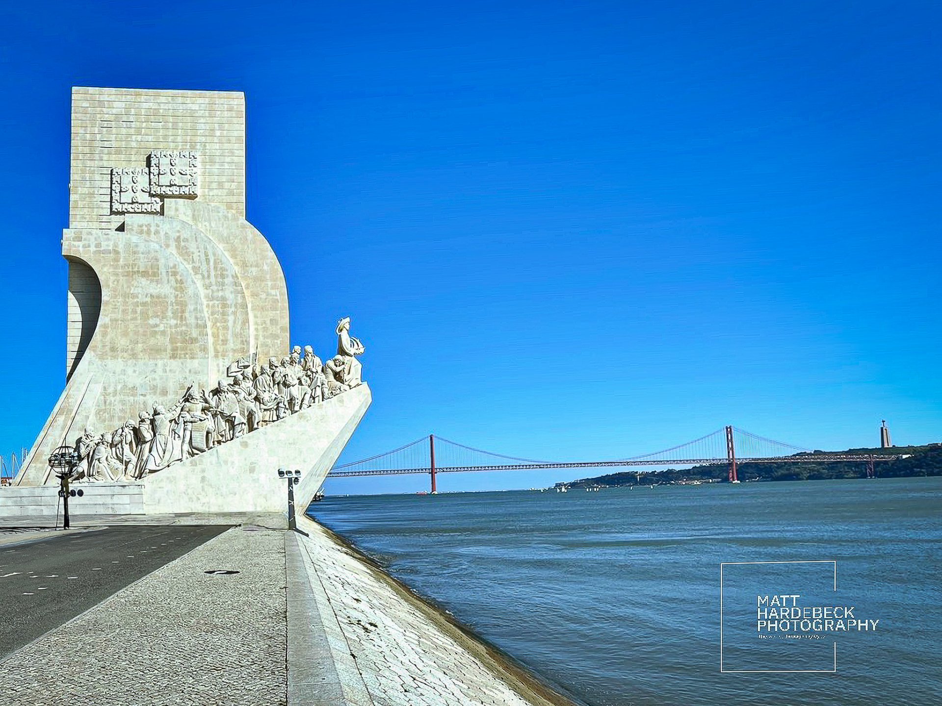 Monument to the Discoveries in Lisbon, Portugal, with statues of historical explorers and the 25 de Abril Bridge in the background.