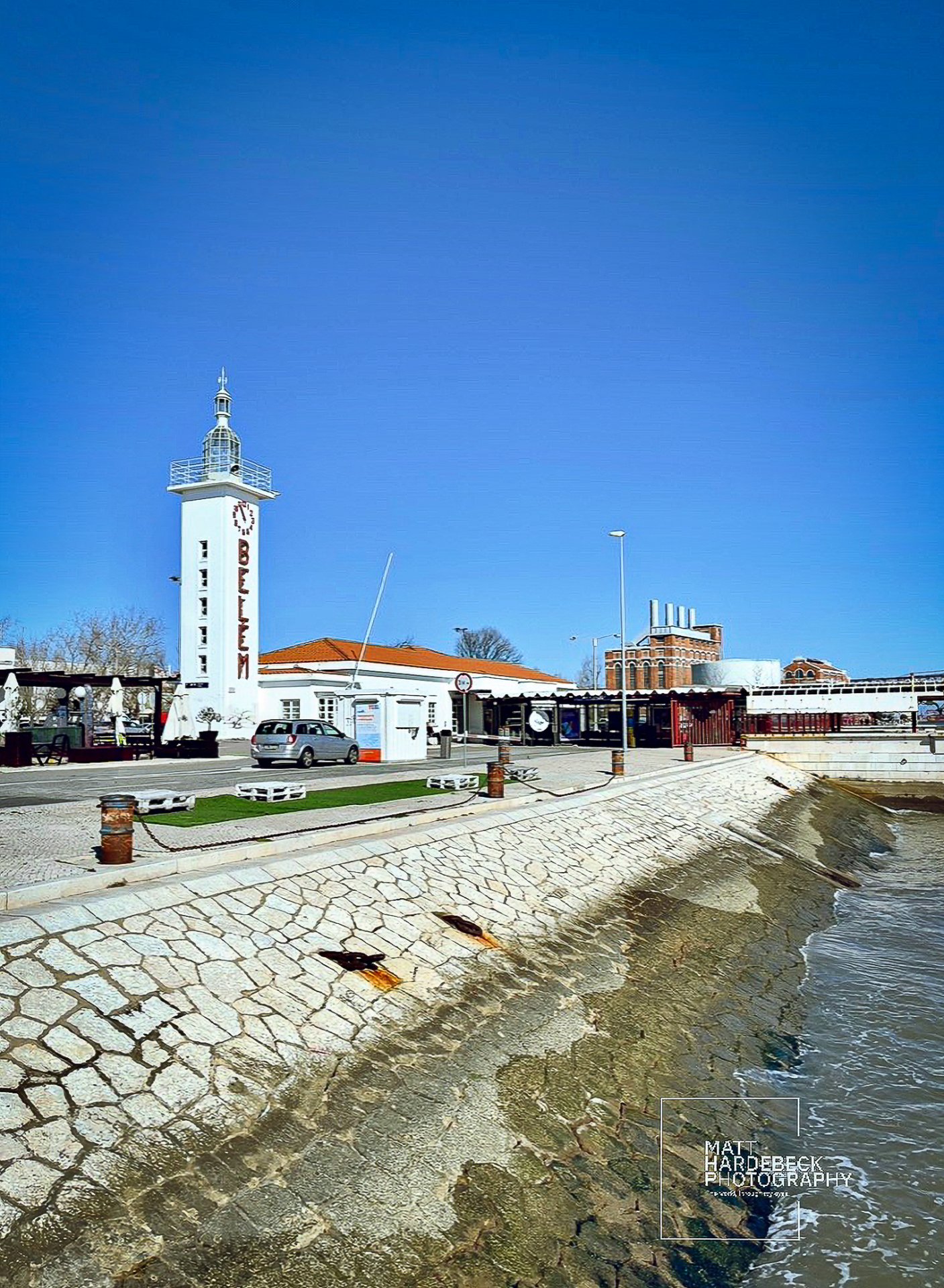 White Belem Lighthouse standing beside the Tagus River under a clear blue sky, with waterfront promenade in the foreground.