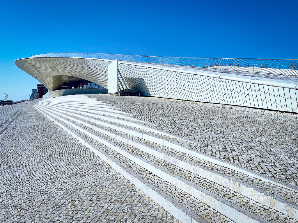 Curved, futuristic white facade of Lisbon’s MAAT museum, with broad stone steps leading up under a cloudless sky.