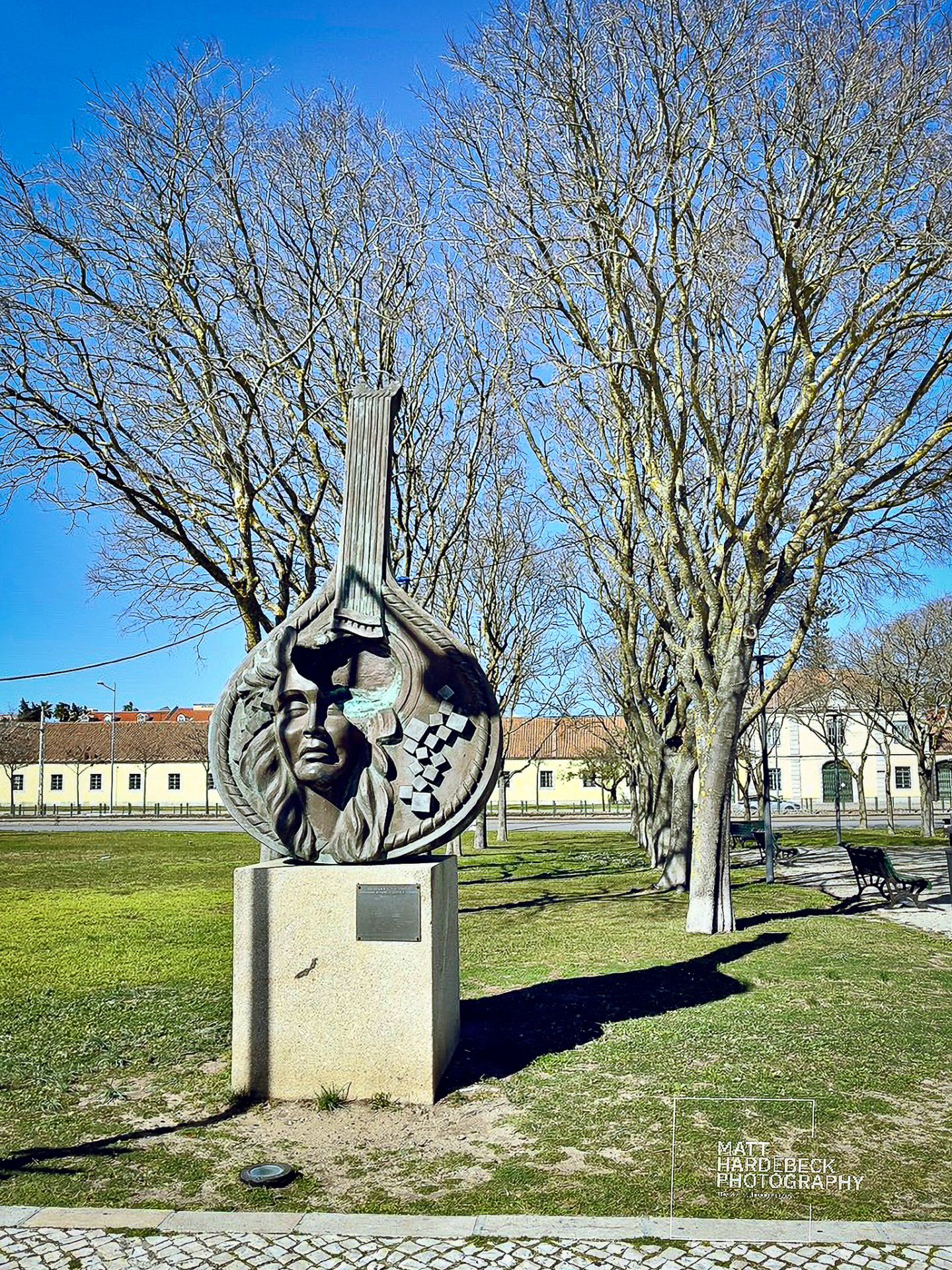 Bronze sculpture featuring a stylized guitar and the face of a fado singer, set against bare winter trees in Lisbon.