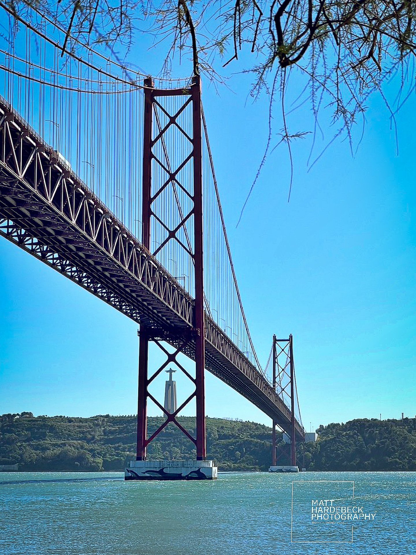 Red suspension bridge Ponte 25 de Abril spanning the Tagus River, with the Cristo Rei statue visible in the distance.