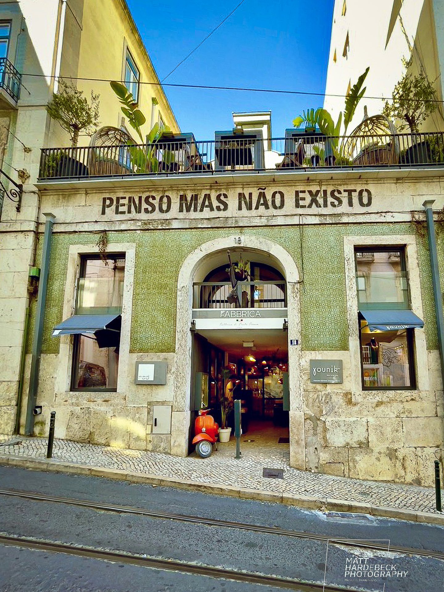 Green-tiled Lisbon building with the phrase "Penso Mas Não Existo" painted above an arched café entrance.