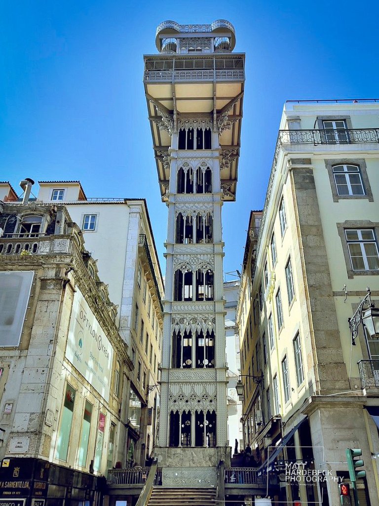 Santa Justa Lift, an ornate neo-Gothic elevator tower in Lisbon, rising between narrow streets under a blue sky.