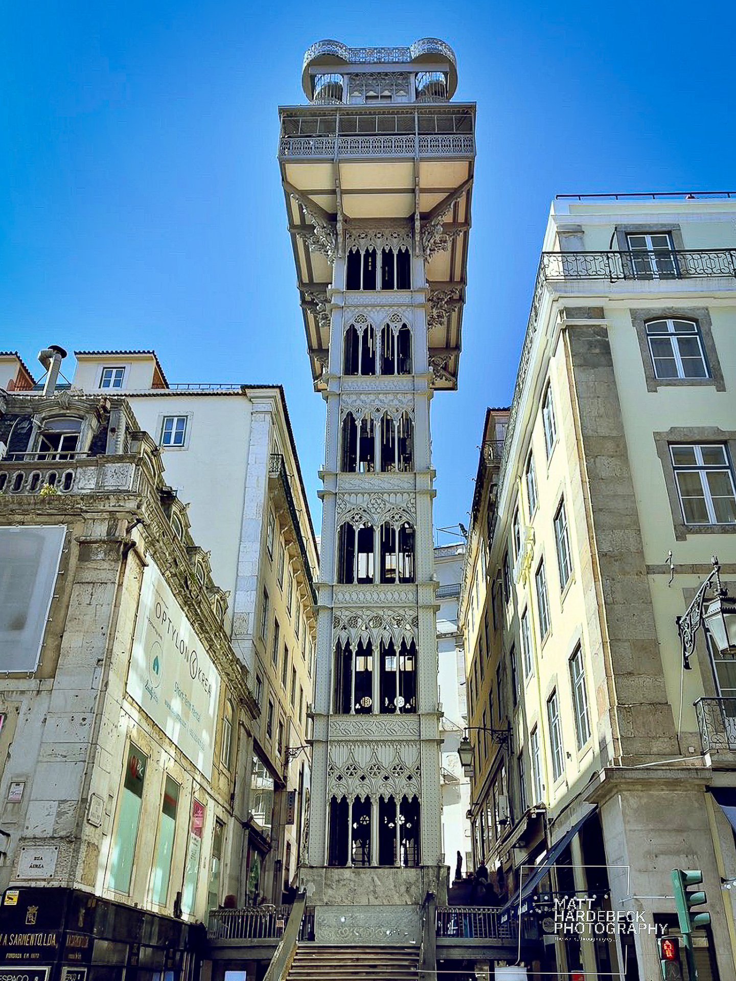 Santa Justa Lift, an ornate neo-Gothic elevator tower in Lisbon, rising between narrow streets under a blue sky.