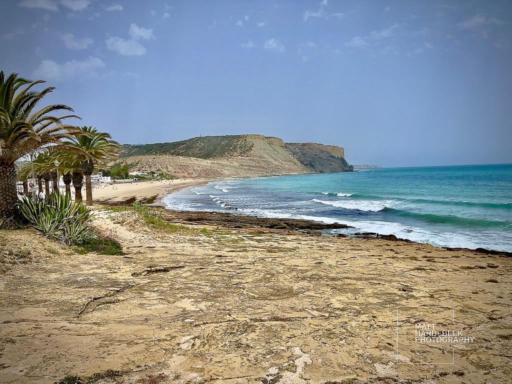 Palm-lined promenade along a sandy beach in Luz, with cliffs rising in the distance.