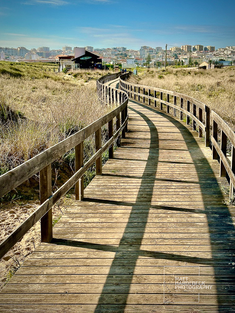 Wooden boardwalk leading through dunes toward Meia Praia, with Lagos skyline in the distance.